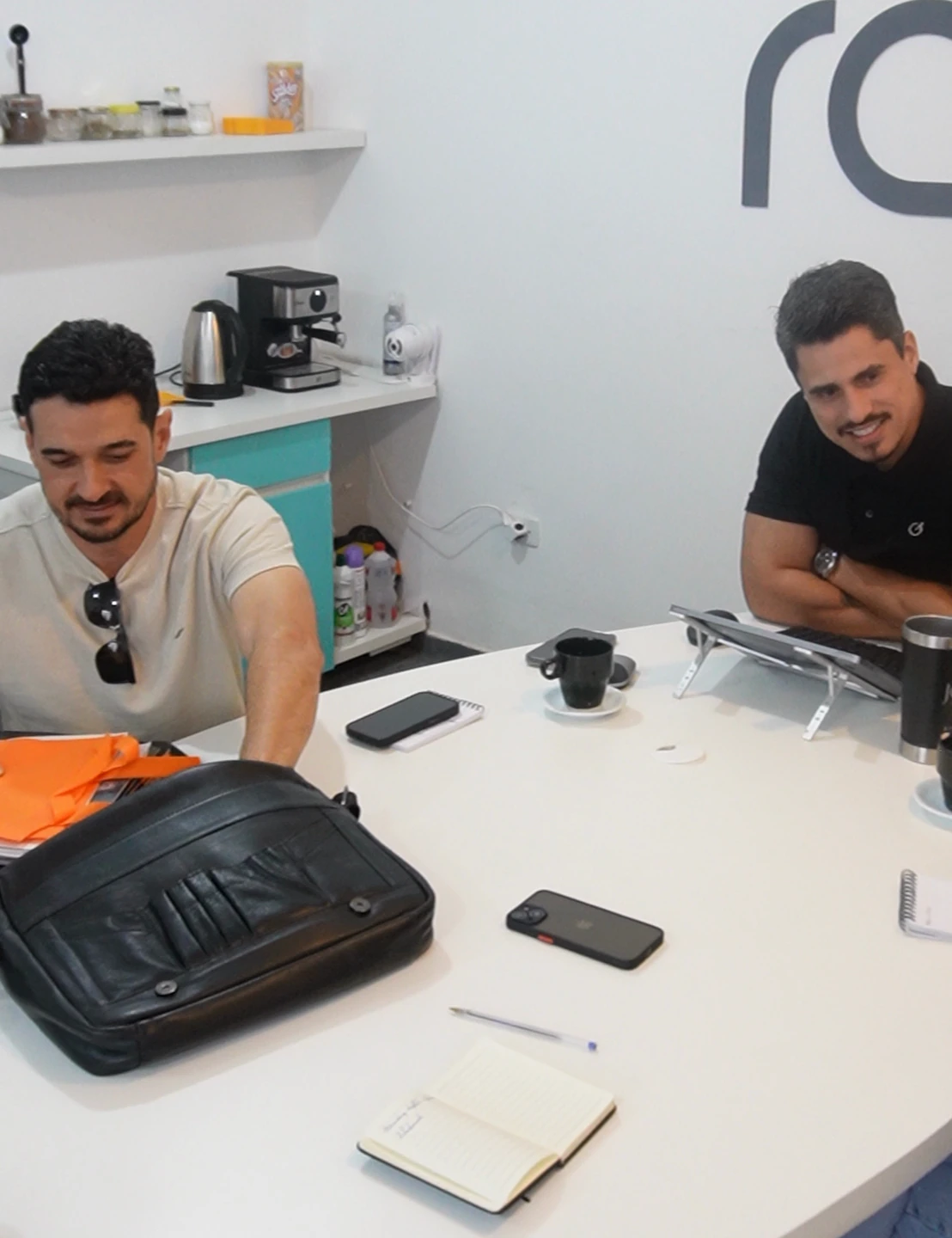 Rafael e outro homem sentados à mesa de reunião, com cadernos e materiais de trabalho.