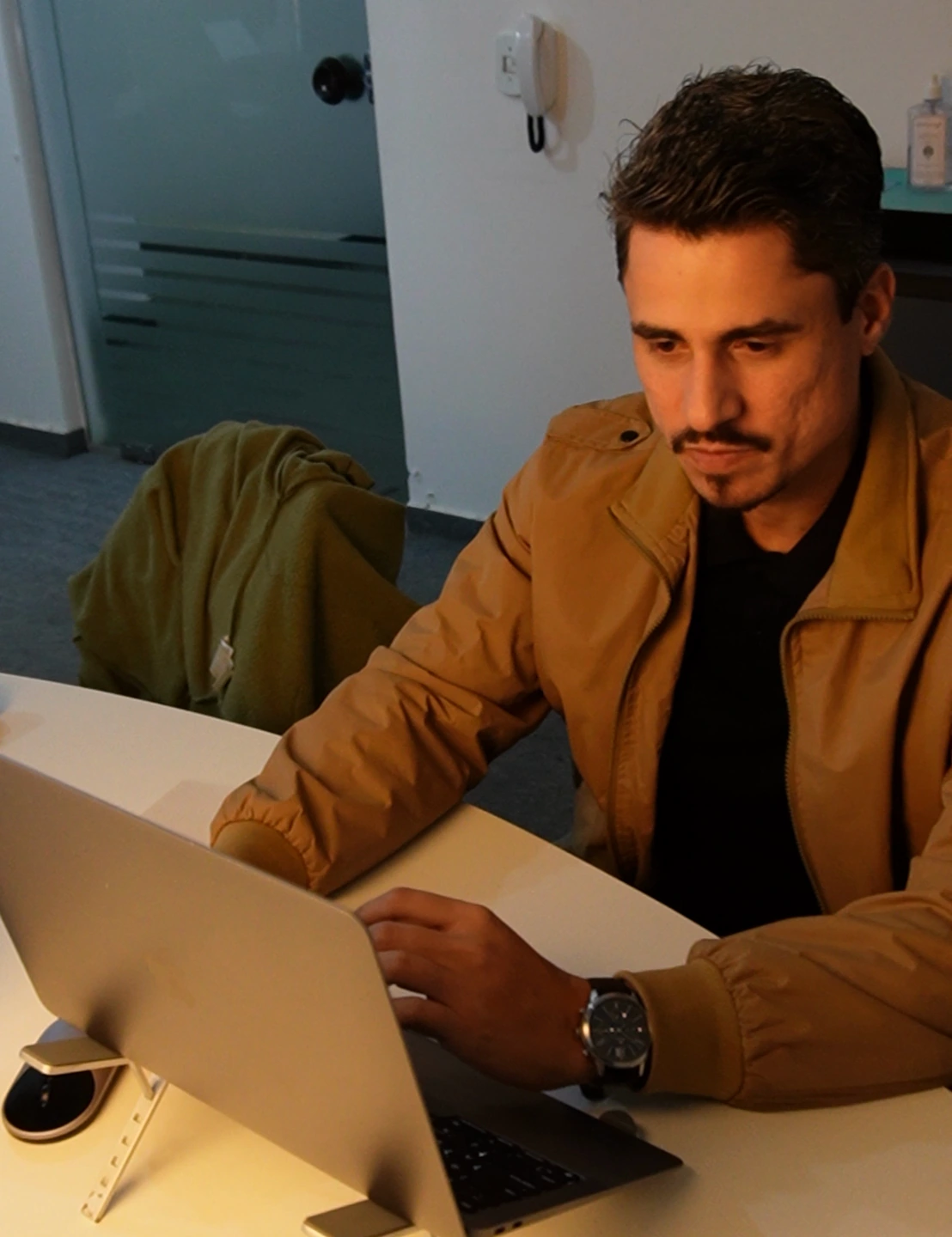 Rafael, de jaqueta ocre, trabalha no notebook apoiado sobre a mesa.
